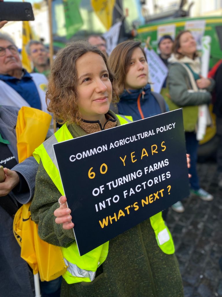 A woman in a demonstration is holding a placard that states 'Common Agricultural Policy, 60 years of turning farms into factories. What's next?"