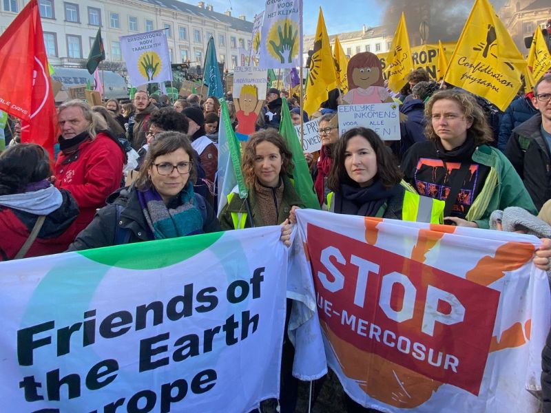 Women holding a flag with Friends of the Earth Europe logo and a flag which reads Stop EU-Mercosur. They are santing in a crowd - a farmers protest - on Place du Luxembourg in Brussels.