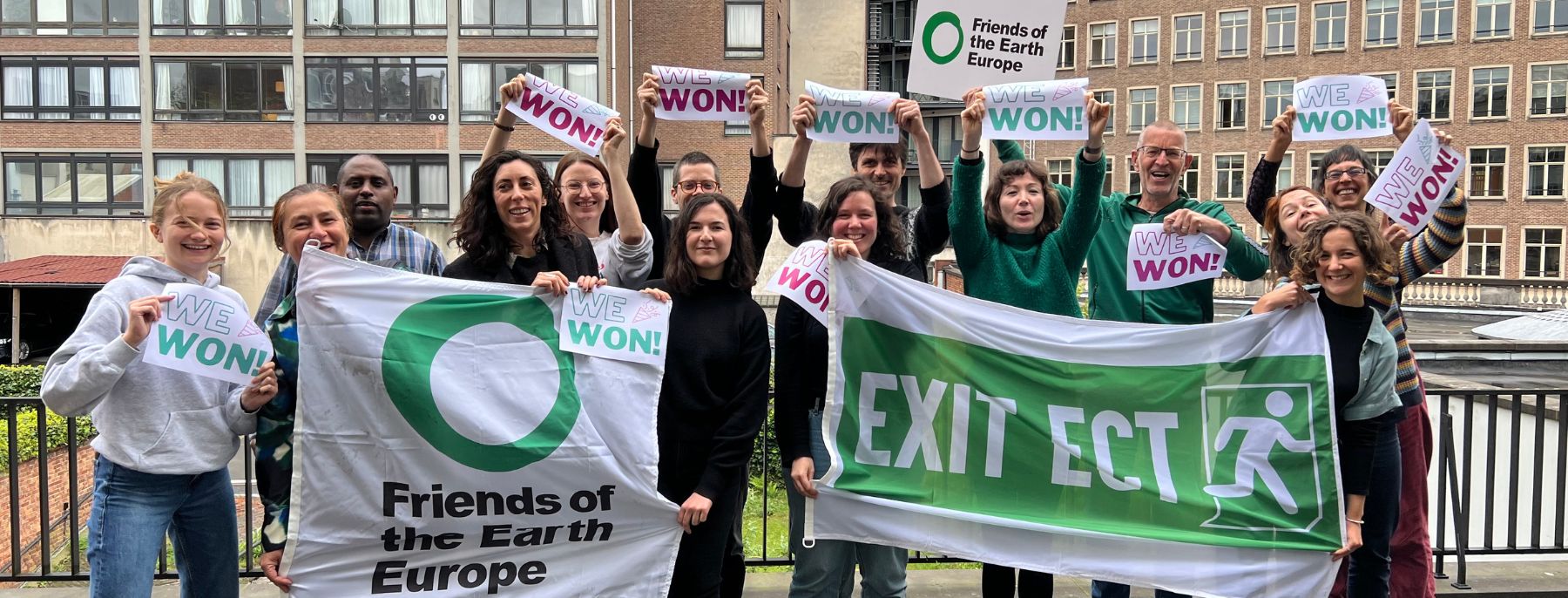 A group of people celebrating outdoor the EU Parliament voting forexiting the ECT,holding signis saying "we won!", a banner saying "Exit ECT" and flags with the Friends of the Earth Europe logo